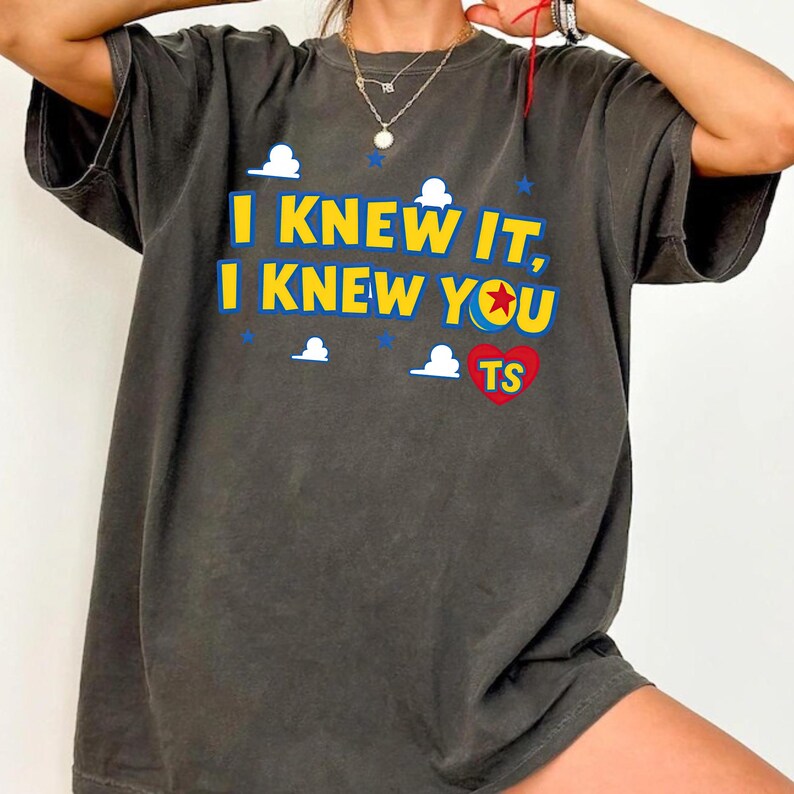 May include: A charcoal gray short-sleeve t-shirt featuring the yellow text I KNEW IT, I KNEW YOU with a red heart containing the letters TS. The design includes small white cloud graphics, blue stars, and a colorful ball icon with a red star.