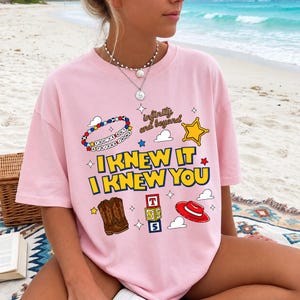 May include: A light pink short-sleeve t-shirt featuring a graphic print with the text I KNEW IT I KNEW YOU, infinity and beyond, and YOU'VE GOT A FRIEND IN ME. The design includes illustrations of a beaded bracelet, a star, cowboy boots, blocks, and a hat.