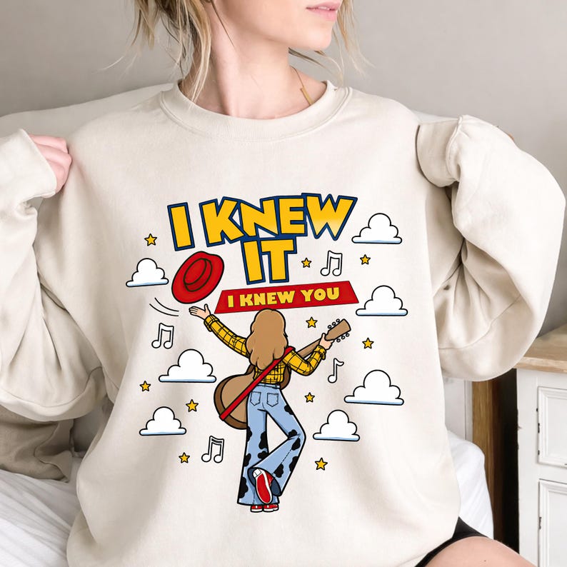 May include: A beige crewneck sweatshirt featuring a graphic illustration of a person with long hair playing a guitar, wearing a yellow plaid shirt and cow-print bell-bottom jeans. The text reads I KNEW IT and I KNEW YOU, with clouds, stars, and musical notes.