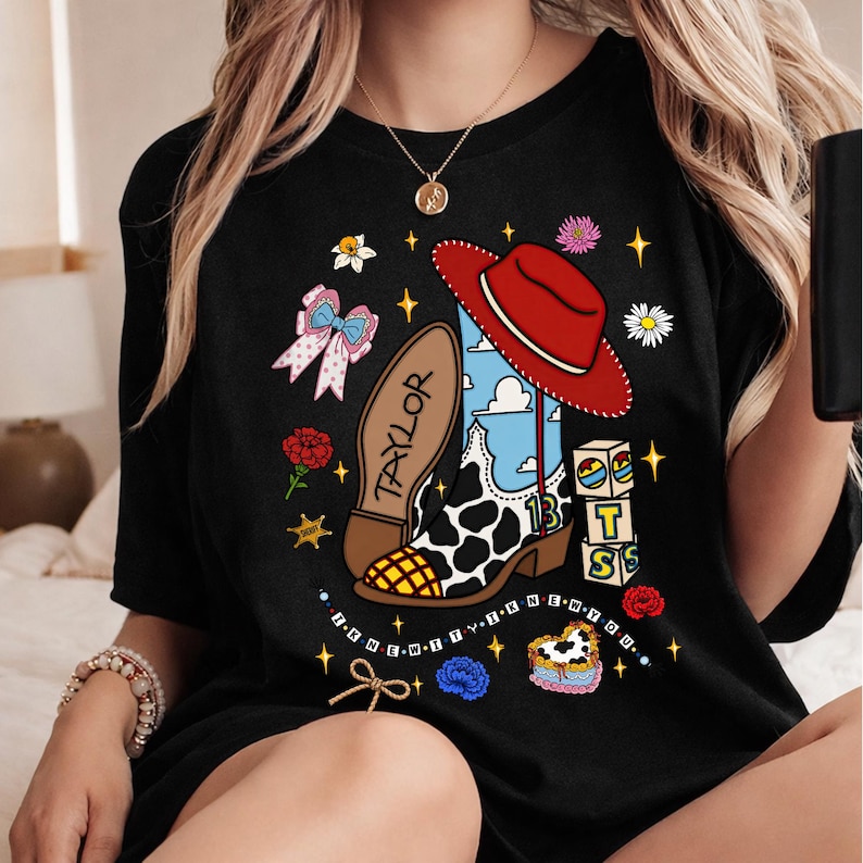Puede incluir: Una camiseta negra con un estampado gr&aacute;fico de una bota de vaquero con estampado de vaca, un sombrero rojo, el nombre TAYLOR, el n&uacute;mero 13 y bloques con las letras T S. El dise&ntilde;o incluye un lazo rosa, flores y el texto I KNEW IT I KNEW YOU.