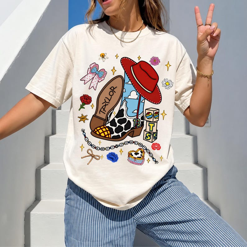 Puede incluir: Una camiseta de manga corta color crema con un estampado gr&aacute;fico de una bota vaquera con manchas de vaca, un sombrero rojo, el nombre Taylor, el n&uacute;mero 13 y varios iconos peque&ntilde;os que incluyen flores, lazos, bloques y cuentas.