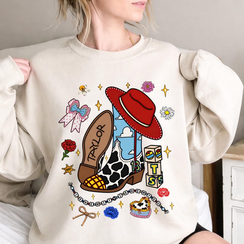 Puede incluir: Una sudadera beige de cuello redondo con un estampado gr&aacute;fico de una bota vaquera con manchas de vaca, un sombrero rojo, el nombre TAYLOR, el n&uacute;mero 13, bloques con las letras T, S, un lazo, flores, estrellas y una cadena de cuentas.
