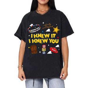 May include: A black t-shirt featuring a graphic print with the yellow text I KNEW IT I KNEW YOU. The design includes a beaded bracelet, a yellow star, brown cowboy boots, a red hat, alphabet blocks, and the text infinity and beyond.