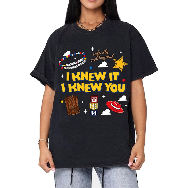 May include: A black t-shirt featuring a graphic print with the yellow text I KNEW IT I KNEW YOU. The design includes a beaded bracelet, a yellow star, brown cowboy boots, a red hat, alphabet blocks, and the text infinity and beyond.