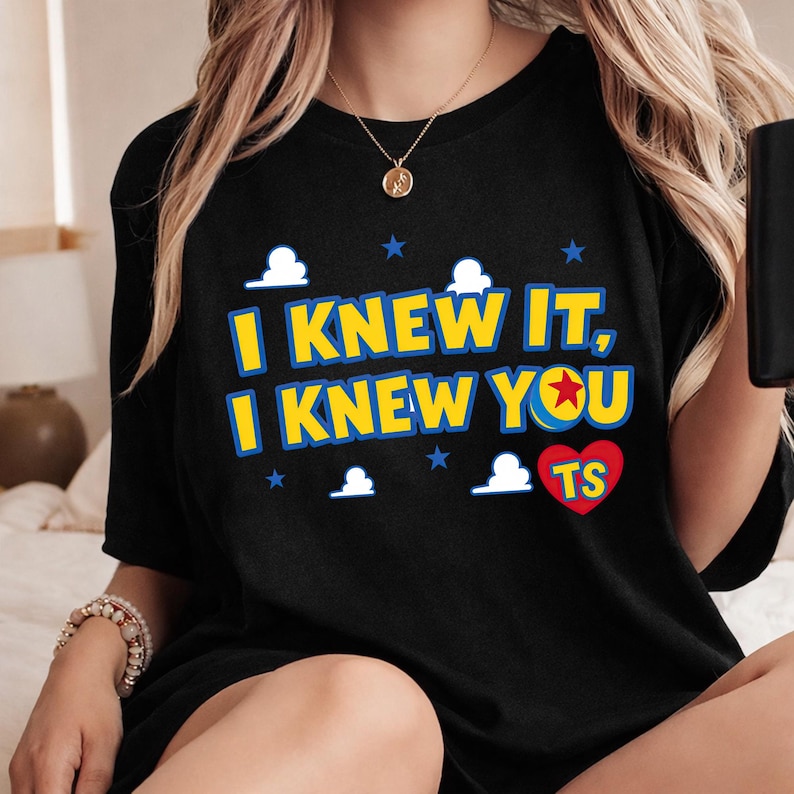 May include: A black short-sleeved t-shirt featuring the yellow text I KNEW IT, I KNEW YOU with a red star inside the letter O. The design includes white cloud shapes, small blue stars, and a red heart containing the yellow letters TS.