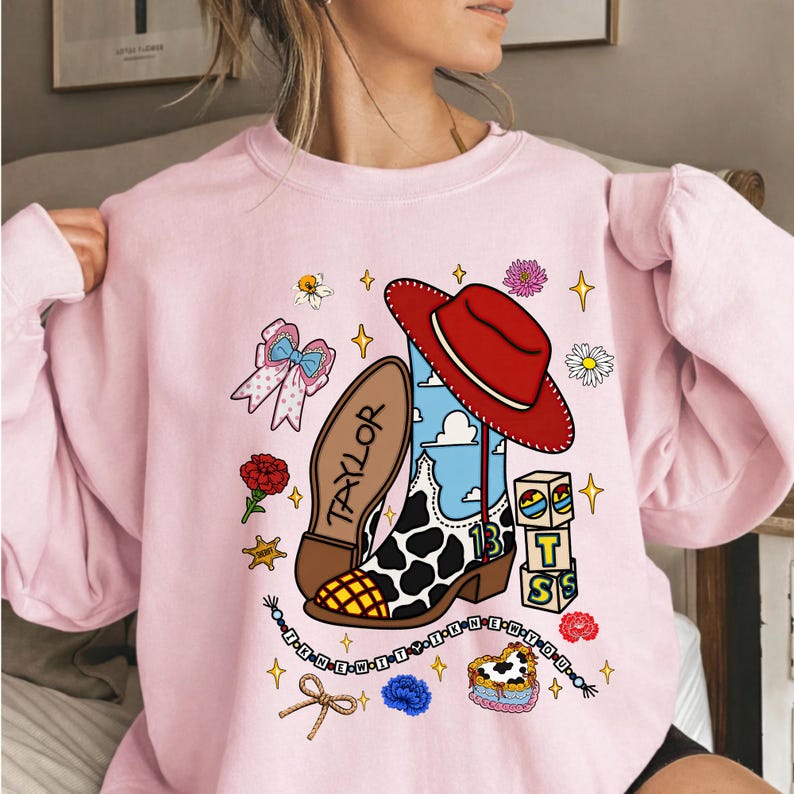 Puede incluir: Una sudadera de cuello redondo rosa claro con un estampado gr&aacute;fico de una bota vaquera con patr&oacute;n de manchas de vaca, un sombrero vaquero rojo, un cielo azul, el nombre Taylor, el n&uacute;mero 13, bloques con letras, flores, un lazo y un collar.