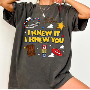 May include: A charcoal gray t-shirt featuring the yellow text I KNEW IT I KNEW YOU, a beaded bracelet with the text YOU'VE GOT A FRIEND IN ME, the text infinity and beyond, a yellow star, brown cowboy boots, alphabet blocks, and a red hat.