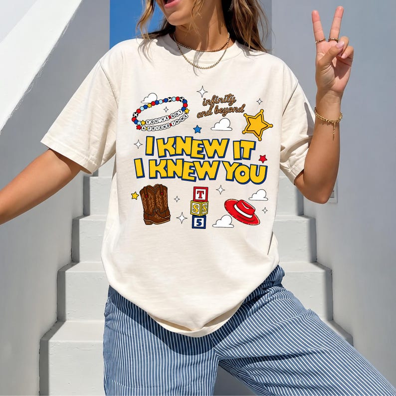 May include: A cream-colored short-sleeve t-shirt featuring the text I KNEW IT I KNEW YOU in bold yellow letters. The graphic includes a beaded bracelet, a sheriff star, brown cowboy boots, alphabet blocks, and a red hat with cloud icons.