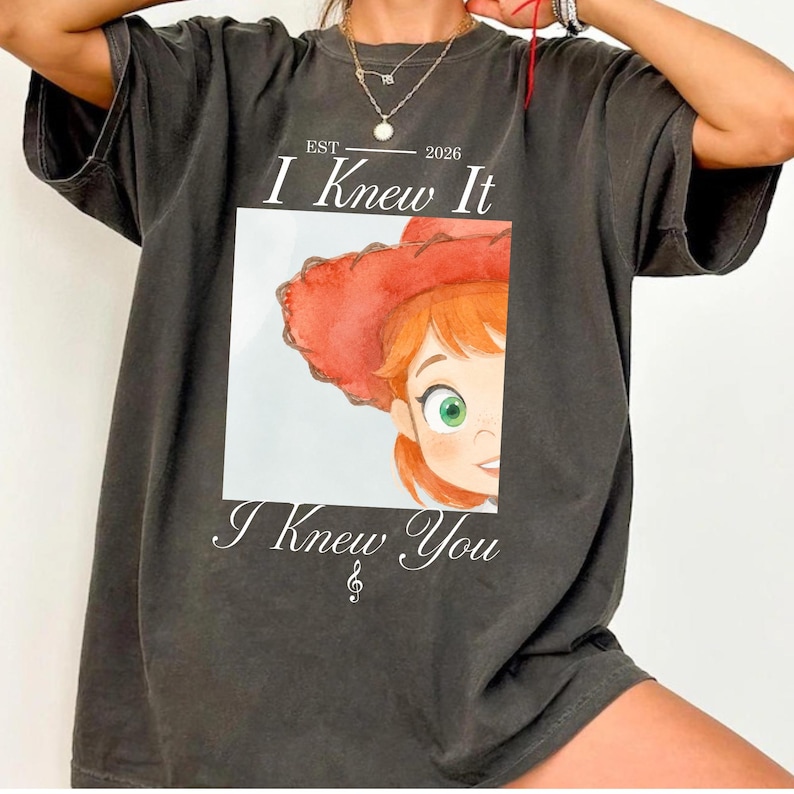 May include: A charcoal gray t-shirt featuring a graphic print of a cartoon character with red hair and a red hat. The text reads EST 2026 I Knew It I Knew You with a small musical note symbol below the text. The shirt has short sleeves and a crew neck.