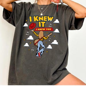 May include: A charcoal gray t-shirt featuring a graphic of a person with long hair playing a guitar, wearing a yellow plaid shirt and cow-print bell-bottom pants. The text reads I KNEW IT and I KNEW YOU, surrounded by white clouds, stars, and music notes.