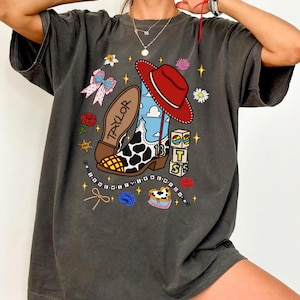 Puede incluir: Una camiseta gris carb&oacute;n de manga corta con un estampado gr&aacute;fico de una bota vaquera con manchas de vaca, un sombrero rojo, una suela con la palabra TAYLOR, bloques con TS, un collar de cuentas, un lazo, flores y estrellas sobre un cielo azul.