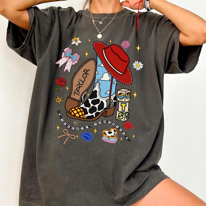 Puede incluir: Una camiseta gris carb&oacute;n de manga corta con un estampado gr&aacute;fico de una bota vaquera con manchas de vaca, un sombrero rojo, una suela con la palabra TAYLOR, bloques con TS, un collar de cuentas, un lazo, flores y estrellas sobre un cielo azul.