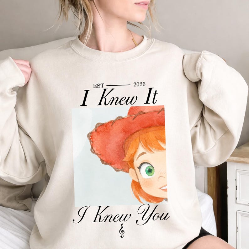 May include: A beige crewneck sweatshirt featuring a graphic print of a cartoon character with red hair and a red hat. The text reads EST 2026 I Knew It I Knew You with a small musical treble clef symbol centered below the text on the lower portion.