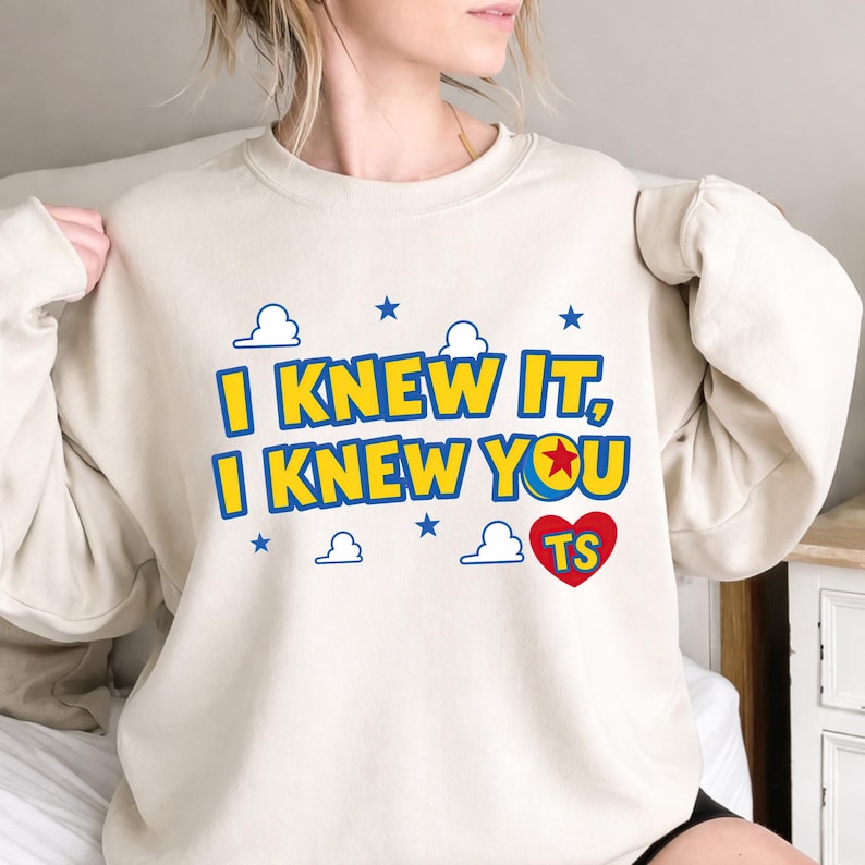 May include: A beige crewneck sweatshirt featuring the yellow text I KNEW IT, I KNEW YOU with a blue outline. The design includes a red heart with the letters TS, a ball with a red star, and small blue clouds and stars printed on the front.