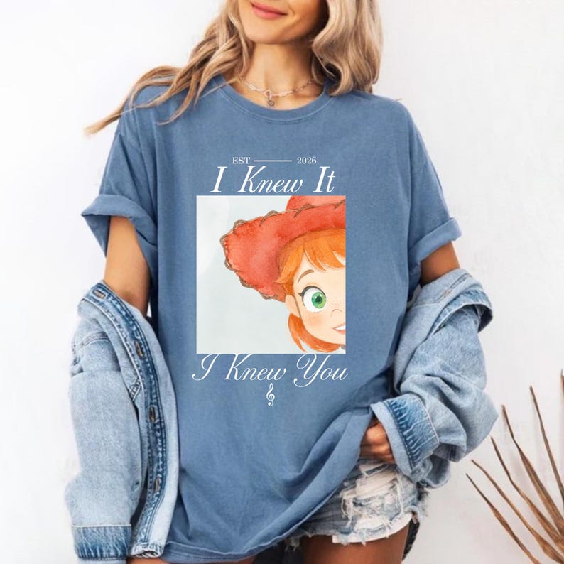May include: A blue short-sleeve t-shirt featuring a graphic of a cartoon character wearing a red cowboy hat. The text "EST 2026 I Knew It I Knew You" is printed in white above and below the square image of the character's face.