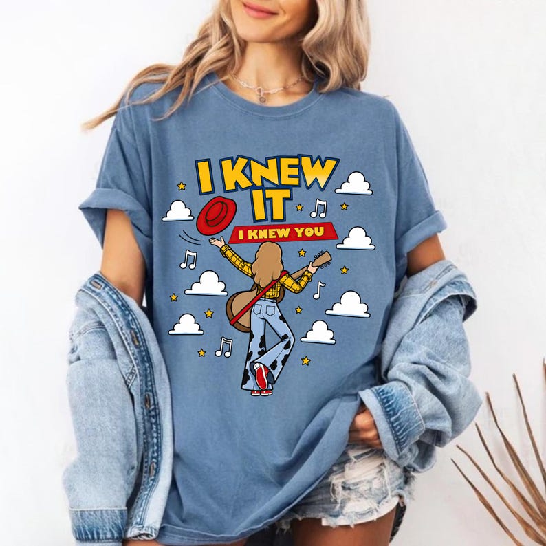 May include: A blue t-shirt featuring a graphic of a person with long hair wearing a yellow plaid shirt and cow-print pants, holding a guitar and a red hat. The text reads I KNEW IT I KNEW YOU, surrounded by white clouds, yellow stars, and musical notes.