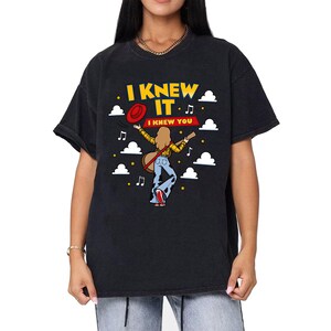 May include: A black short-sleeved t-shirt featuring a graphic of a person with long hair wearing a yellow plaid shirt and cow-print bell-bottoms, holding a guitar and a red hat. The text reads I KNEW IT and I KNEW YOU, surrounded by clouds and stars.