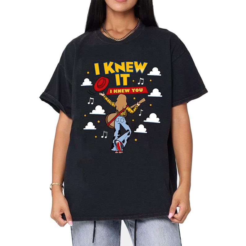 May include: A black short-sleeved t-shirt featuring a graphic of a person with long hair wearing a yellow plaid shirt and cow-print bell-bottoms, holding a guitar and a red hat. The text reads I KNEW IT and I KNEW YOU, surrounded by clouds and stars.
