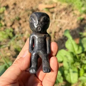 May include: A small, dark grey figurine of a humanoid figure with large eyes and a small mouth. The figurine is carved from a smooth, possibly stone material. The figure is standing with its arms at its sides.