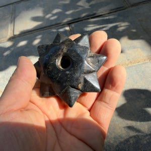 May include: A black, star-shaped object with pointed edges and a central hole. The object is held in a hand, showcasing its dark color and geometric design. The star-shaped item appears to be made of a solid, possibly stone-like material.