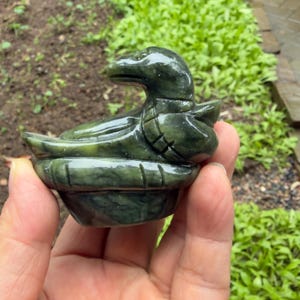 May include: A dark green jade duck figurine, hand-carved with intricate details. The duck rests in a carved bowl-shaped base. The jade has a polished finish, and the duck's form is clearly defined. The figurine is a decorative item.