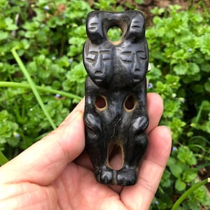 May include: A carved black stone figurine with a stacked design. The top features two faces with a circular hole above them. Below, two more faces are carved, and the bottom has a large opening. The figurine is held in a hand.
