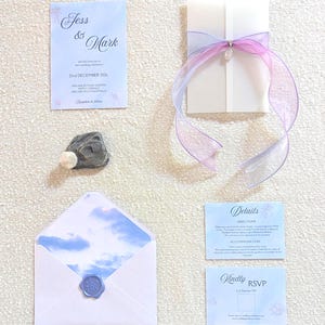 Könnte beinhalten: Hochzeitseinladungs-Set in Hellblau und Weiß. Enthält eine Einladungskarte, eine RSVP-Karte, eine Detailkarte und einen Umschlag mit Wachssiegel. Eine weiße Karte ist mit einem lila Band gebunden. Ein Stein und eine Muschel sind ebenfalls vorhanden.