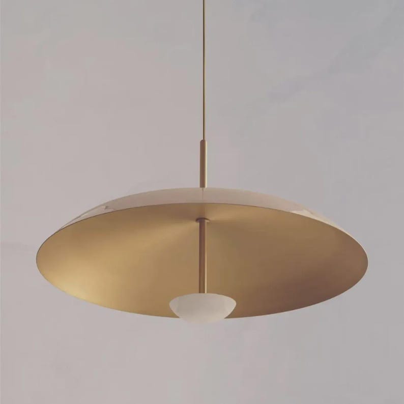 May include: A modern, gold-toned pendant light fixture. The light has a large, circular shade with a downward-facing, frosted glass bowl. The fixture is suspended from a thin rod.