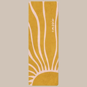May include: A rectangular yoga mat featuring a mustard yellow background with a light pink sunburst pattern radiating from the bottom center. The name "cassie" is printed in a white cursive script font vertically along the right side of the mat.