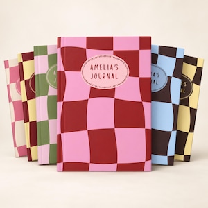 May include: A collection of six hardcover journals arranged in a fan shape, each featuring a large checkered pattern in various color combinations. The central journal is pink and dark red with an oval label reading AMELIA'S JOURNAL in red text.
