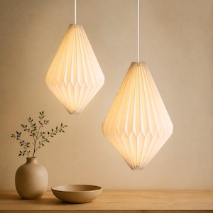 Op de afbeelding: Twee gebroken witte, ruitvormige hanglampen met een geplooide, papierachtige textuur. Elke lamp hangt aan een dun wit snoer en verspreidt een warme, zachte gloed. De kappen hebben een geometrisch, gevouwen ontwerp dat taps toeloopt.