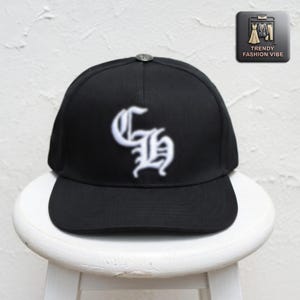 May include: Black baseball cap with a white embroidered gothic-style "GH" logo. The cap has a curved brim and a small silver button on top. The cap is sitting on a white stool. The Trendy Fashion Vibe logo is in the upper right corner.