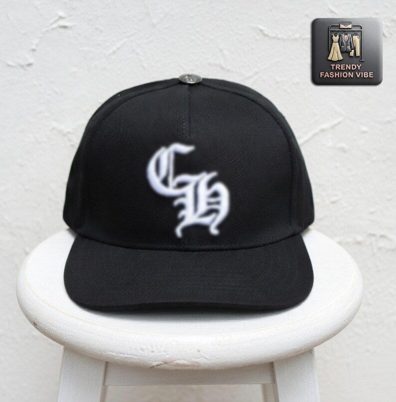 May include: Black baseball cap with a white embroidered gothic-style "GH" logo. The cap has a curved brim and a small silver button on top. The cap is sitting on a white stool. The Trendy Fashion Vibe logo is in the upper right corner.