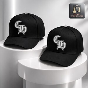 May include: Two black baseball caps with a curved brim are displayed. Each cap features a white, stylized letter design on the front. The caps are on a white surface. A small silver button is on top of each cap. The caps are part of the Trendy Fashion Vibe collection.