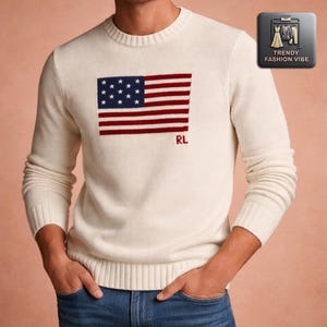 May include: Cream-colored sweater featuring a stylized American flag design in navy blue and red. The flag is embroidered on the front, with the initials "RL" in red below. The sweater has a crew neck and long sleeves.