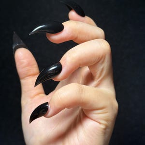 May include: A set of long, pointed, glossy black press-on nails applied to the fingers. The nails feature a sharp, curved stiletto shape with a smooth, reflective finish that covers the natural nail bed, extending outward to a distinct, tapered point.