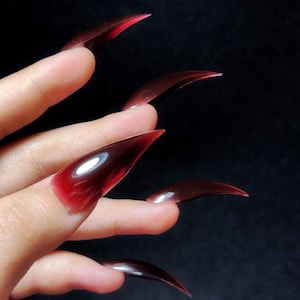 May include: A hand with long, pointed, translucent red nails. The nails have a gradient effect, transitioning from a deep red at the tips to a lighter shade near the base. The background is a dark, solid color.