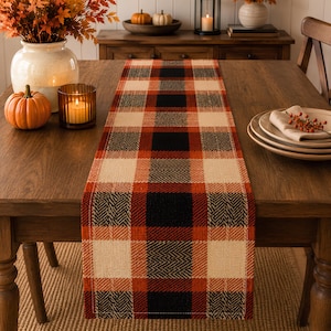 Peut inclure: Un chemin de table rectangulaire présentant un motif à carreaux tissé dans des tons de noir, orange brûlé et crème. Le tissu affiche une texture à chevrons visible sur tout le motif, créant un aspect structuré sur la table en bois.