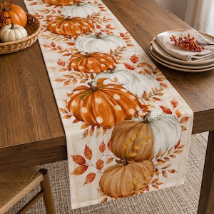 Peut inclure: Un chemin de table rectangulaire orné d'un motif répétitif de citrouilles orange et blanches à pois, entremêlées de feuilles d'automne orange et brunes sur un fond à carreaux beige clair, posé à plat sur une table à manger en bois.
