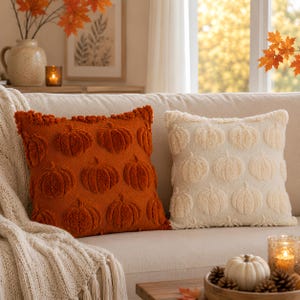 Puede incluir: Dos cojines cuadrados con un patrón de calabazas en relieve y con textura. Un cojín es de color naranja quemado intenso y el otro es de color blanco crema. Ambos cojines tienen una superficie texturizada y afelpada sobre un sofá.