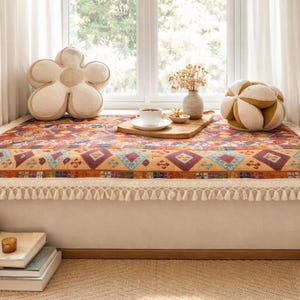 May include: A window seat with a patterned textile, a flower-shaped cushion, and a spherical cushion. A wooden tray holds a cup of coffee and a small dish. A vase with dried flowers sits nearby. The seat has a fringed edge.