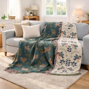 May include: A teal and beige throw blanket with a bird and floral pattern draped over a gray sofa. The blanket has a fringed edge and is paired with beige and white throw pillows. The room is well-lit with natural light.