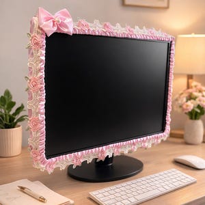 May include: A pink monitor cover with a decorative lace and floral trim. A large pink bow with a pearl accent is at the top. The cover is on a black computer monitor, with a keyboard and mouse in the foreground.
