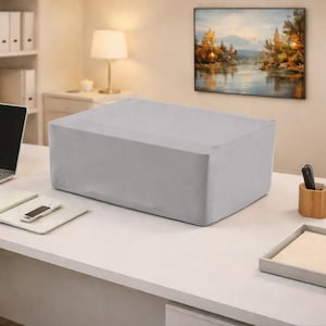 May include: A light gray rectangular object, possibly a piece of furniture or a large box, is covered with a fitted fabric. The object sits on a white desk, with a laptop, phone, and accessories nearby. A painting hangs on the wall.