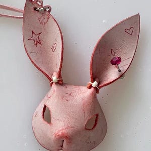 Puede incluir: Un colgante de cuero rosa con forma de conejo con una cara cosida, ojos recortados y una nariz cosida. El colgante tiene orejas largas con diseños de estrellas y corazones grabados y un cordón rosa. Una pequeña gema rosa está unida a una oreja.