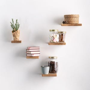 Op de afbeelding: Houten zwevende planken met diverse items. De planken bevatten een potplant, boeken, potten met marshmallows en koffiebonen, een klein mandje en een blauwe beker. De items zijn geplaatst tegen een witte muur.