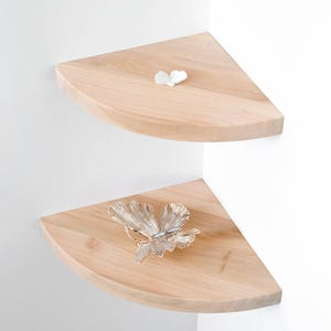 May include: Two light-coloured wooden corner shelves mounted on a white wall. The top shelf holds a small white butterfly decoration, while the lower shelf displays a silver butterfly ornament. The shelves are semi-circular.