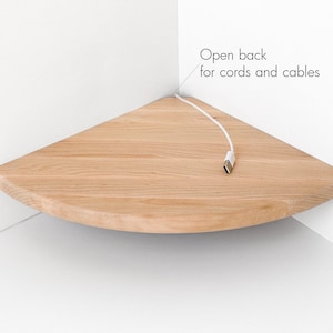 Puede incluir: Una estantería de esquina de madera clara con un borde frontal redondeado. Un cable USB blanco está enchufado a la estantería. El texto "Open back for cords and cables" es visible.