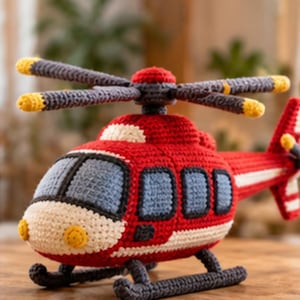 May include: A handcrafted crochet helicopter toy featuring a vibrant red body with white accents, grey windows, and dark grey landing skids. The top rotor blades are grey with yellow tips, and the tail rotor is also grey with yellow accents.
