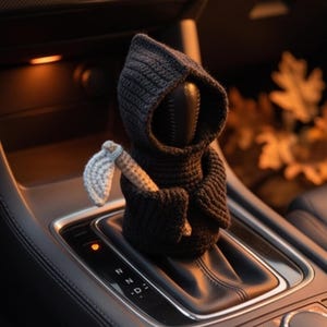 May include: A black crocheted cover for a car gear shift knob, shaped like a hooded figure. The figure wears a dark cloak with a hood and holds a small, light gray crocheted scythe in its hand, positioned over the vehicle's gear selector console.